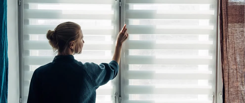 Frau öffnet moderne Doppelrollos am Fenster und reguliert das Tageslicht im Raum.